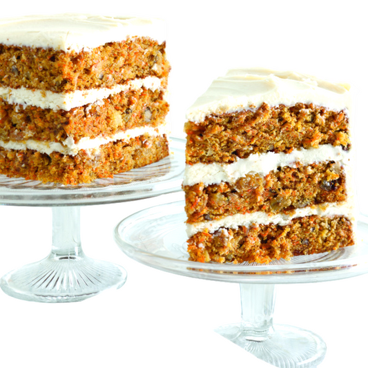 Carrot cake with white frosting on a clear cake stand with a slice removed, on a light wooden surface.
